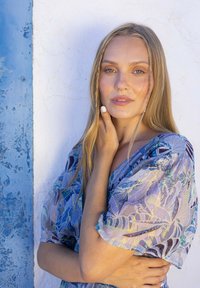 Une robe fluide et légère présente un motif abstrait bleu et violet avec des manches bouffantes, devant un mur blanc et une porte bleue.
