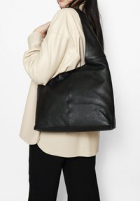 Grote zwarte lederen tote bag met een gestructureerd oppervlak, met een slouchy ontwerp en een enkele schouderband, gedragen tegen een lichte top.