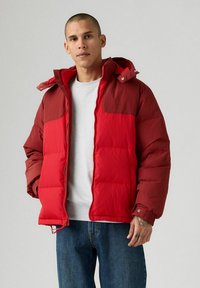 Uomo che indossa un giubbotto imbottito rosso con cappuccio, una camicia grigia chiara e jeans blu, in piedi davanti a uno sfondo grigio uniforme.