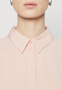 Camicia col colore rosa chiaro con chiusura a bottoni. Presenta una texture liscia e piccoli bottoni trasparenti. Design semplice con colletto classico.