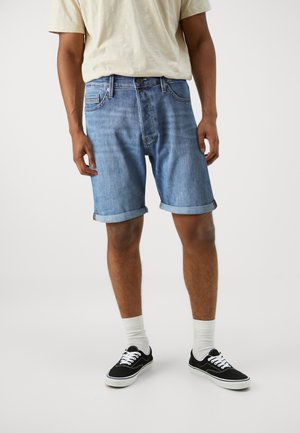 Shorts de denim en azul claro con dobladillos enrollados, que presentan un diseño clásico de cinco bolsillos y un cierre de botón. Combinados con zapatillas de lona negras.