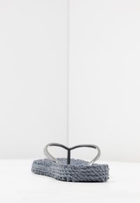 Grå flip-flop med en flettet stofstruktur og sølvfarvede Y-formede stropper. Flad sål, minimalistisk design, sat mod en hvid baggrund.
