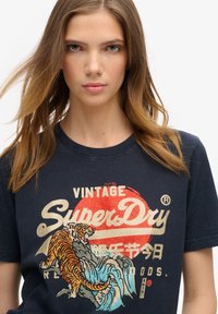 T-shirt en coton bleu marine avec un graphique vibrant d'un tigre et de vagues, avec le texte "Vintage SuperDry" en blanc et des accents dorés.