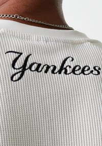 Off-white textured fabric t-shirt with "Yankees" embroidered in black cursive on the back, featuring a round neckline and ribbed pattern.