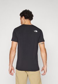 Crna majica kratkih rukava sa zakrivljenim ramenskim dijelom, bijelim logotipom The North Face na gornjem dijelu leđa i ravnim rubom.