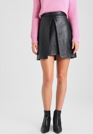 Vrouw draagt een zwarte mini-rok van imitatieleren met een split aan de voorkant, een roze gebreide trui en zwarte enkellaarsjes, staand tegen een effen achtergrond.