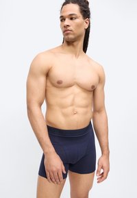 Homme musclé aux cheveux longs en boxer bleu foncé, debout devant un fond clair uni, regardant vers sa gauche.