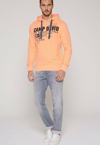 Felpa arancione con dettagli blu navy e grafiche sul davanti, abbinata a jeans grigi chiari e sneaker bianche. Il tessuto sembra morbido e informale.
