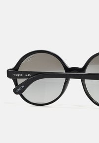 Óculos de sol redondos pretos com lentes degradê, apresentando uma armação larga e braços de templo ousados marcados com "Vogue MBB" e "Londres."