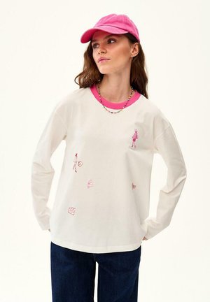 Kobieta w białej koszuli z długim rękawem z różowym kołnierzykiem oraz małymi różowymi nadrukami graficznymi, ciemne jeansy, różowa czapka i kolorowy naszyjnik z koralików.