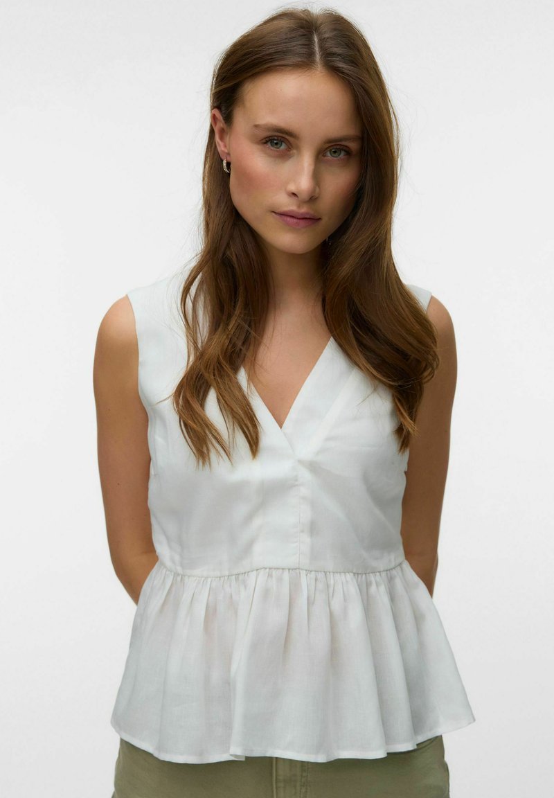 Femme aux longs cheveux bruns, portant de petites boucles d'oreilles anneaux et un haut blanc sans manches à col en V avec volant, faisant face à l'avant sur un fond uni.