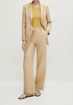 Kobieta ubrana w beżową, dopasowaną marynarkę i szerokie spodnie, z musztardowożółtą bluzką i beżowymi sandałami, stojąca na jednolitym tle.