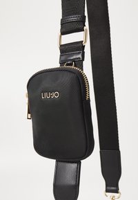 Must must crossbody kott, millel on kuldne riistvara, lukuga sulgemine ja logo detail. Valmistatud siledast materjalist, lame, kompaktne kuju ja reguleeritav rihm.