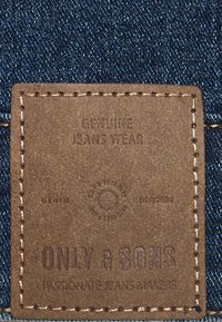Brauner Lederpatch mit prägtem Text auf dunkelblauem Denim. Enthält "ONLY & SONS" und "GENUINE JEANS WEAR" mit genähten Kanten.
