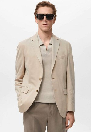 Blazer beige con dos botones, un bolsillo en el pecho, combinado con un polo de punto beige claro. El modelo lleva gafas de sol oscuras y está de pie contra un fondo blanco.