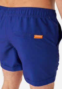 Pantaloni da nuoto blu con vita elasticizzata, dotati di tasca posteriore e etichetta "OPPOSUITS" arancione. Tessuto liscio con una vestibilità aderente.
