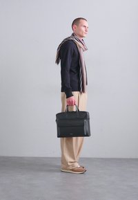 Black leather briefcase with dual top handles, front pocket, rectangular shape, smooth texture. Worn by a figure in a striped scarf and beige trousers.