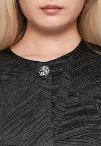 Lähedane pilt naise kaelast ja huultest, kes kannab musta tekstuuri rõivast, millel on kinnitatud üks läikiv hõbedane dekoratiivne nupp.