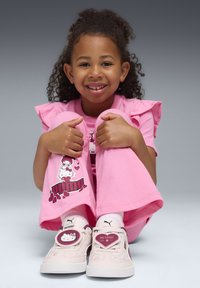 Sneakers Puma rosa con design di Hello Kitty, dotati di un'etichetta a forma di cuore e accenti marrone chiaro. Il modello indossa una maglietta rosa con maniche a volant e pantaloni coordinati.