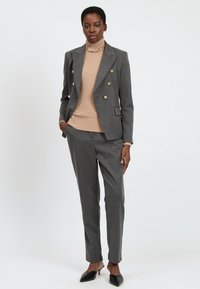 Donna in piedi che indossa un blazer grigio doppiopetto con bottoni dorati, pantaloni coordinati, dolcevita beige a coste e tacchi a punta neri.