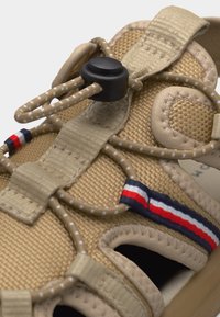 Gros plan sur une sandale beige avec des lacets bungee ajustables, un bouton-poussoir noir et un détail de bride en tissu rouge, blanc et bleu.