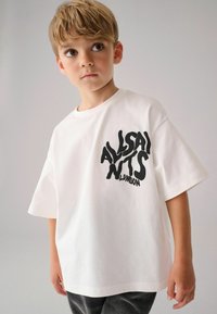 Bílé oversized tričko s černým nápisem "ALLSAINTS LONDON" v vlnitém stylu. Bavlněný materiál, krátké rukávy, kulatý výstřih, jednoduchý design.