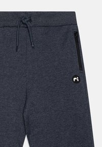 Mørk grå joggebukser laget av myk stoff, med snøring i livet og en glidel�åslomme på siden. Inkluderer en svart brodert logo.