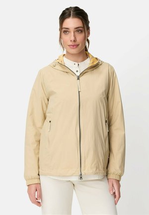 Frau, die eine beige Reißverschluss-Kapuzenjacke über einem weißen Hemd und einer hellen Hose trägt, steht vor einem schlichten hellen Hintergrund.