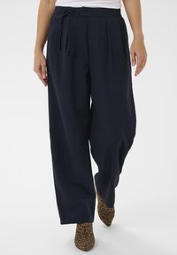 Woman wearing loose navy pants with a drawstring waist and leopard-print pointed boots, walking forward on a white background.