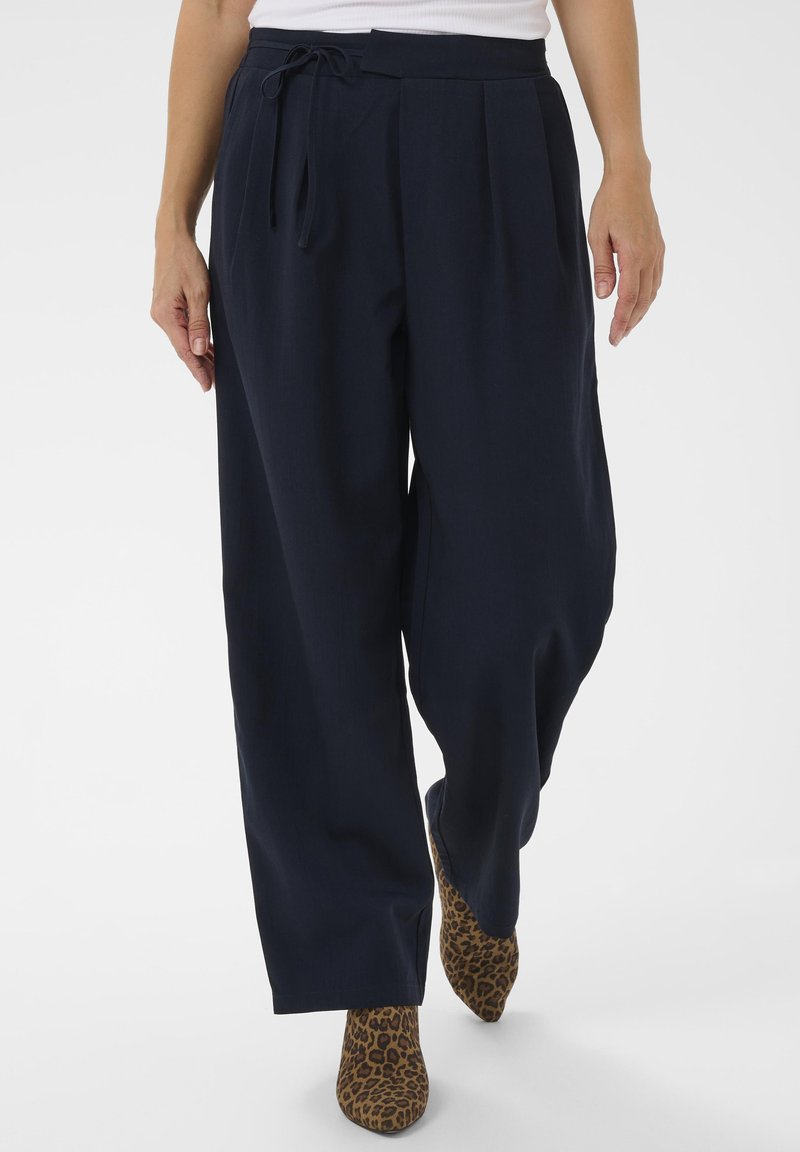 Ženska v širokih temno modrih hlačah z vrvico v pasu in s škornji z leopardjim vzorcem, ki hodi naprej na beli podlagi.