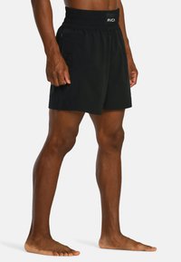 Man in zwarte RVCA sportshorts staat blootsvoets tegen een plain achtergrond.