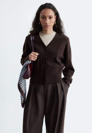 Femme aux longs cheveux bouclés porte un cardigan boutonné marron foncé sur une chemise claire et un pantalon ample foncé, tenant un petit sac avec une écharpe.