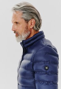 Granatowa kurtka puchowa z wysokim kołnierzem, z poziomym przeszyciem, zamek błyskawiczny oraz okrągła czarna naszywka z logo na rękawie.