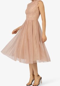 Femme portant une robe sans manches en dentelle rose poudré avec une jupe en tulle, associée à des sandales à talons hauts avec des brides transparentes.