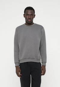 Sivá mikina s okrúhlym výstrihom, dlhými rukávmi a rebrovanými manžetami. Tkanina je hladká s jemnou textúrou. Skombinovaná s čiernymi nohavicami.