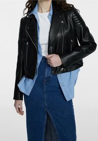 Giubbotto da motociclista in pelle nera con pannelli spalle testurizzati, tasche con zip, indossato sopra una camicia di jeans azzurri chiari e una gonna di jeans blu scuro.