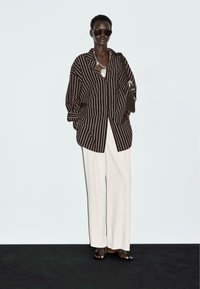 Gestreepte bruin-witte oversized shirt, witte wijde broek en donkere zonnebril. Accessoires omvatten een zwarte tas en gelaagde kettingen.