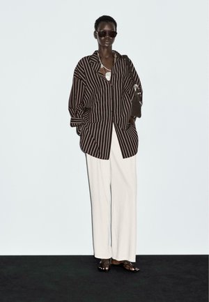 Camicia oversize a righe marroni e bianche, pantaloni bianchi a gamba larga e occhiali da sole scuri. Accessori includono una borsa nera e collane a strati.