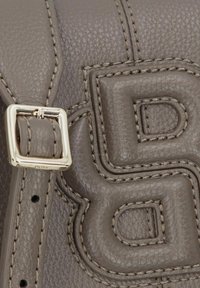 Primer plano de un bolso de cuero texturizado color topo con detalle de la letra "B" cosida y una hebilla rectangular plateada en la correa.