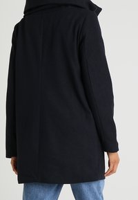 Persona che indossa un cappotto nero di lunghezza intermedia con un collo ampio, in piedi con la schiena rivolta verso la fotocamera, abbinato a jeans azzurri chiari.