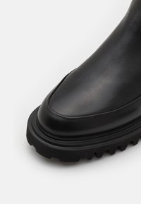 Stivale alla caviglia in pelle nera con punta arrotondata, suola in gomma spessa con un motivo frastagliato, texture liscia e design minimalista.