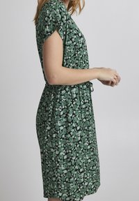 Vihreä kukkamekko, jossa on lyhyet hihat, vyötärömuotoilu ja löysä istuvuus. Mekossa on tumma tausta, jossa on eri sävyisiä vihreitä ja valkoisia kukkia.