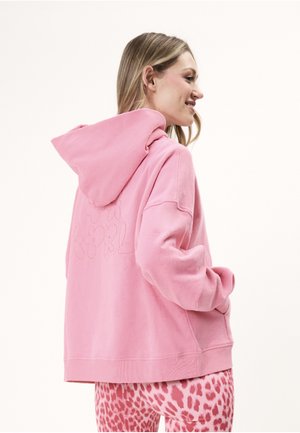 Kvinde iført en pink hættetrøje med prægede bogstaver på bagsiden og pink leopardprint leggings, smilende og kigger til siden.