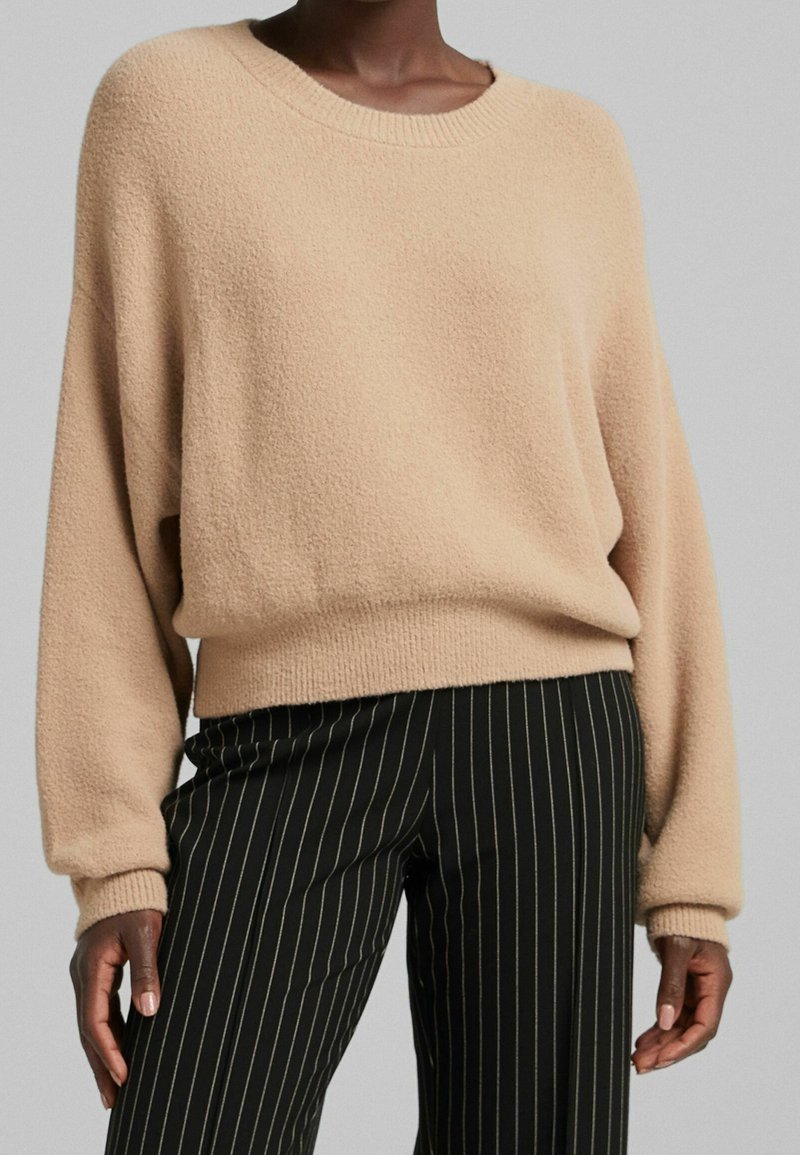 Personne portant un pull beige texturé à manches longues et un pantalon noir à fines rayures verticales blanches, debout devant un fond uni.