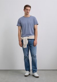 Ein junger Mann steht, trägt ein blaues Kurzarmhemd, helle Jeans, weiße Sneaker und einen beigen Pullover, der um seine Taille gebunden ist.