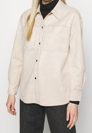 Osoba ubrana w beżową, zapinaną na guziki kurtkę na czarny golf oraz czarne jeansy, stojąca na jednolitym tle.