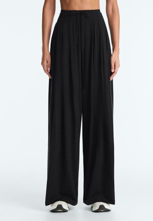 Žena v čiernych širokých voľných nohaviciach s nastaviteľným pásom a bielych teniskách, stojaca pred jednoduchým svetlým pozadím.