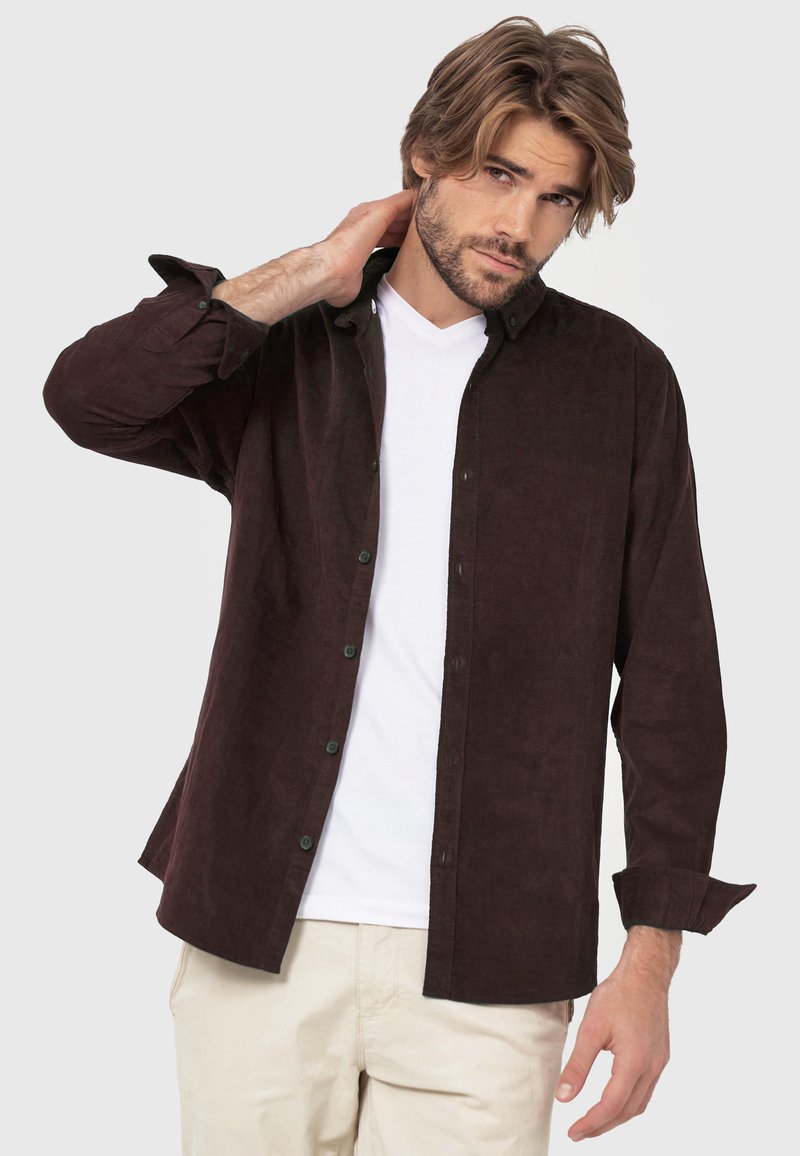 Braune Corduroy-Button-Up-Hemd mit lockerer Passform, langen Ärmeln und grünen Knöpfen. Trägt man über einem weißen Rundhals-T-Shirt.
