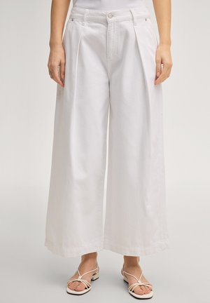 Vrouw draagt witte, high-waisted, wijd uitlopende broek met voorplooi en knoopsluiting, gecombineerd met witte sandalen met bandjes.
