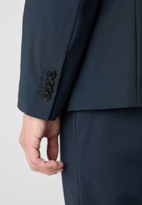 Manica di giacca blu scuro con quattro bottoni neri e dettagli di cucitura, superficie in tessuto liscia e vestibilità sartoriale.